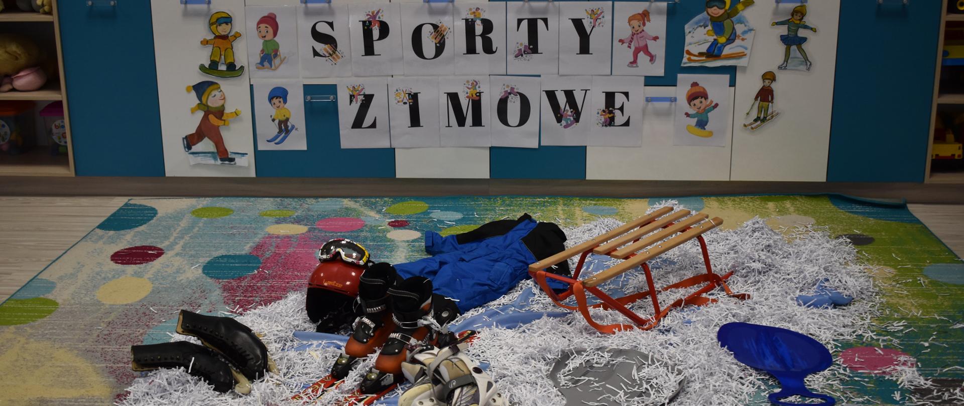 Zdjęcie przedstawia żłobkową salę. W tle na regałe przyklejony jest napis Sporty zimowe oraz plansze je przedstawiające. Na dywanie rozsypane jest biełe konfetti imitujące śnieg, na którym leżą łyżwy, narty i sanki.