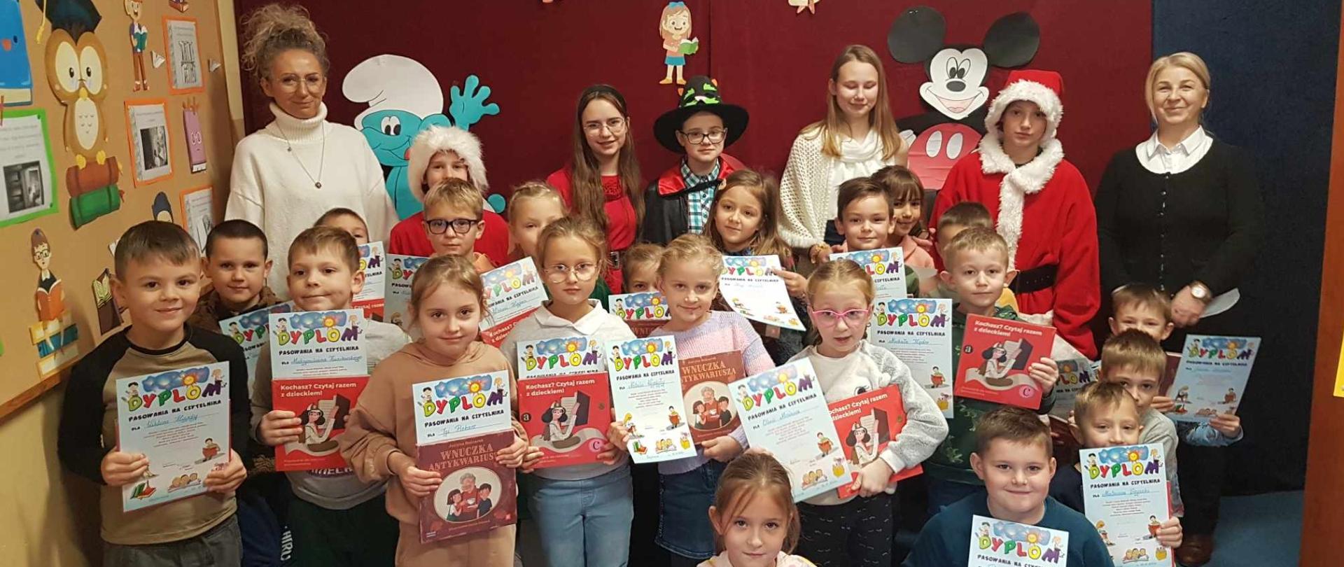 Uczniowie w bibliotece szkolnej, po ceremonii pasowania, ustawieni wspólnie z Panią Dyrektor oraz Panią Bibliotekarką na tle pięknej bajkowej dekoracji, z książkami i dyplomami w dłoniach.