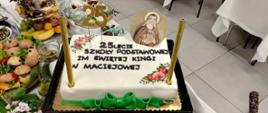 Tort z napisem "25lecie Szkoły Podstawowej im. Świętej Kingi w Maciejowej", ozdobiony w rogach kwiatami i okrągłym wizerunkiem Świętej Kingi