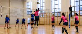 Fotografia z rozgrywek na hali sportowej meczu siatkówki, po lewej stronie drużyna w niebieskich koszulkach po prawej w różowych. Na pierwszym planie dwie zawodniczki z przeciwnych drużyn walczą o piłkę. Z lewej strony na podeście przy siatce stoi sędzia.