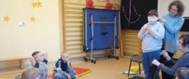 Podczas zajęć - prowadząca zajęcia zawiązała oczy jednemu dziecku celem przeprowadzenia ćwiczeń. Pozostałe dzieci siedzą na matach