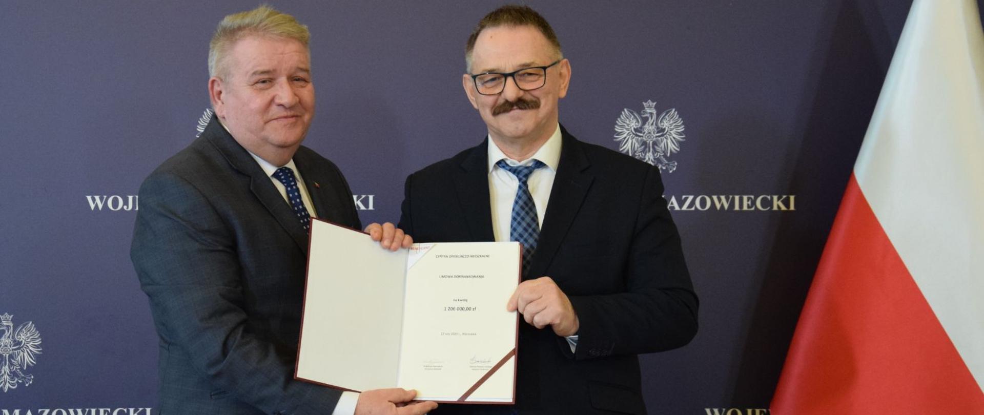 Wojewoda Mazowiecki i Starosta Lipski trzymają podpisaną umowę. W tle granatowa ścianka z logo Wojewody Mazowieckiego. Z prawej strony Flaga Polski.