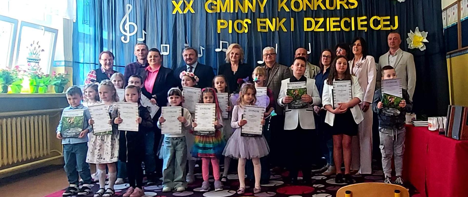 Grupa dzieci i dorosłych w eleganckich strojach. Dzieci przed sobą trzymają dyplomy. Za grupą znajduje się granatowe tło a na nim napis XX Gminny Konkurs Piosenki Dziecięcej. Po lewej stronie duże okno, na parapecie stoją doniczki z kwiatami. 