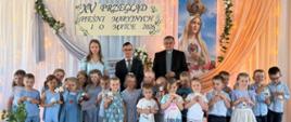 Liczna grupa przedszkolaków ubrana w błękitne i denimowe stroje pozuje na scenie wspólnie z opiekunką i dwoma mężczyznami. Dzieci trzymają w rączkach małe, pluszowe misie.