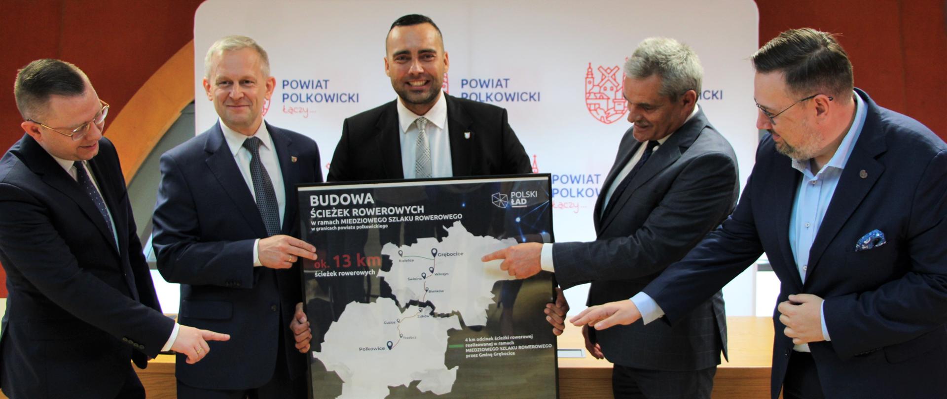 Władze powiatu polkowickiego oraz zaproszeni goście trzymają planszę z mapą ścieżek rowerowych i wskazują na nią palcem 