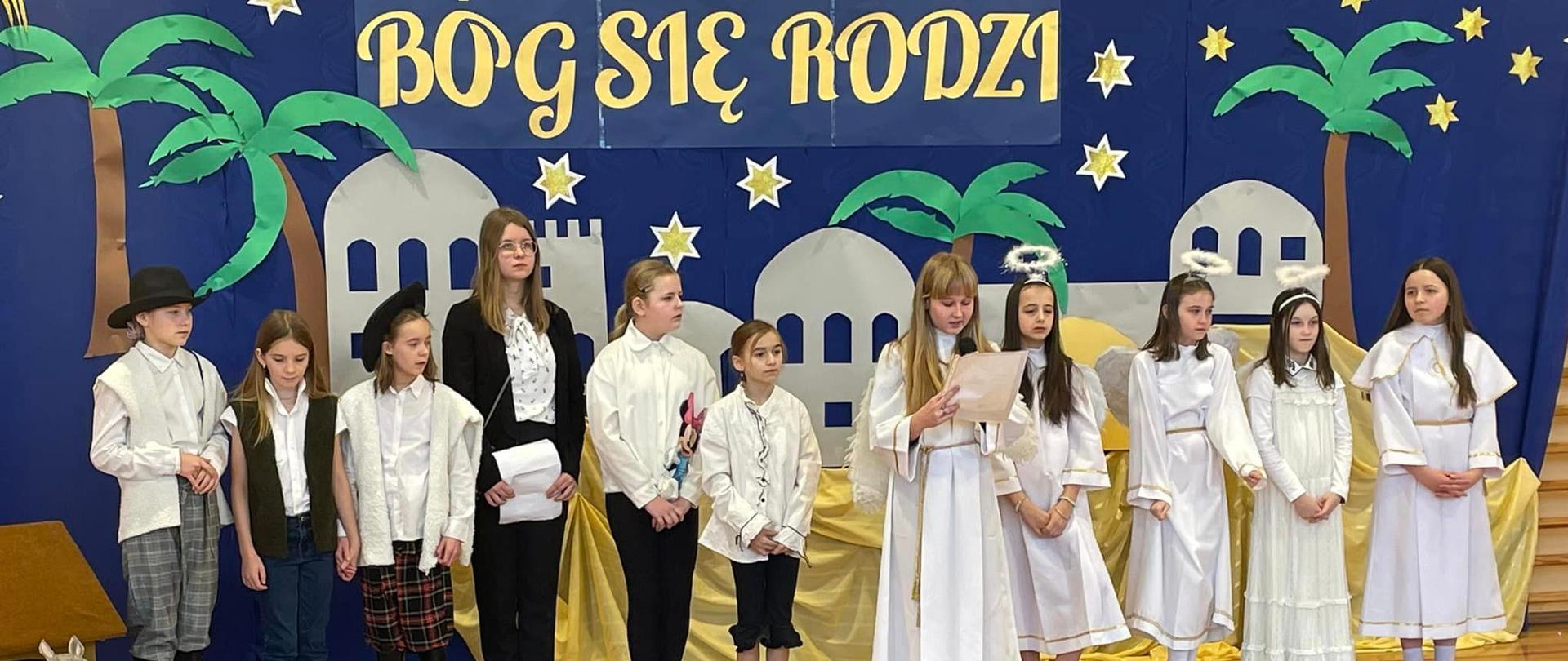Na tle dekoracji z napisem Bóg się rodzi stoją uczniowie występujący w jasełkach, przebrani w stroje świąteczne, widoczne są również głowy widzów oglądających występ
