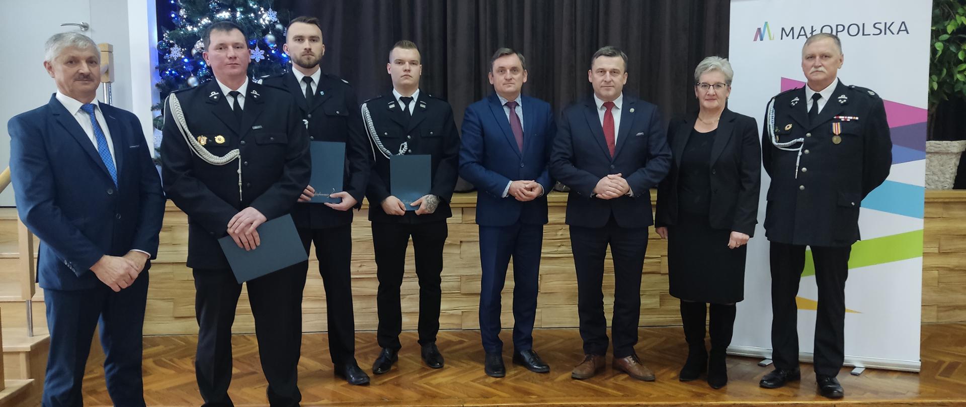 Grupa osób stoi w rzędzie na sali. Wśród nich funkcjonariusze w galowych mundurach oraz przedstawiciele władz w garniturach. Kilku funkcjonariuszy trzyma teczki z dokumentami. W tle choinka oraz roll-up z logo Małopolski.