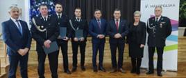 Grupa osób stoi w rzędzie na sali. Wśród nich funkcjonariusze w galowych mundurach oraz przedstawiciele władz w garniturach. Kilku funkcjonariuszy trzyma teczki z dokumentami. W tle choinka oraz roll-up z logo Małopolski.