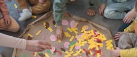 Zdjęcie przedstawia duża papierową pizzę wraz z kolorowymi papierowymi dodatkami tj żółte paski, zielone listki, różowe koła, czerwone półokręgi i brązowe pieczarki. Dookoła pizzy siedzą dzieci ale widać tylko nóżki dłonie, nie widać twarzy. Dzieci dotykają poszczególnych dodatków do pizzy i rozkładają je po tekturowym cieście. 
