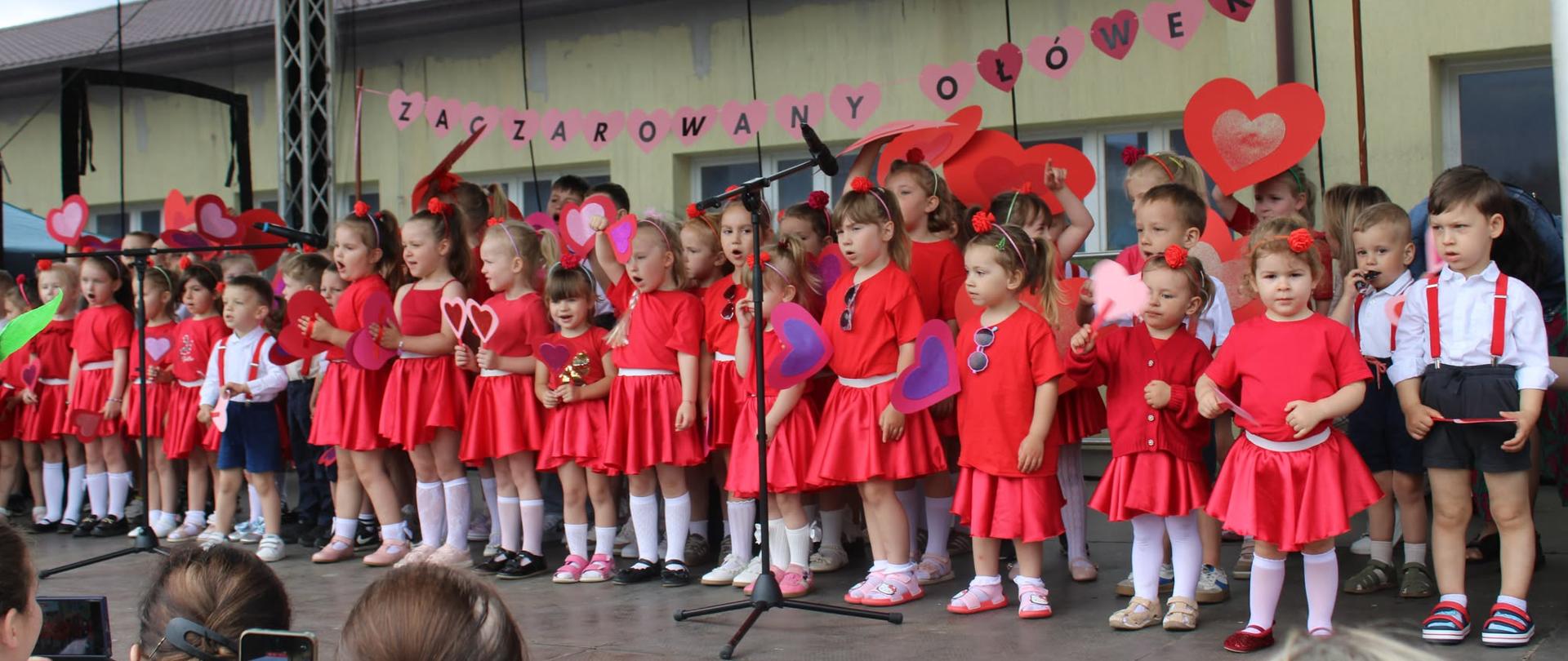 Duża grupa małych dzieci (około 40) w czerwonych sukienkach i białych strojach z czerwonymi szelkami wykonuje spektakl na scenie plenerowej. Dzieci trzymają kolorowe serca z papieru. W tle baner z napisem "Zaczarowany Ołówek". Publiczność fotografuje wydarzenie.