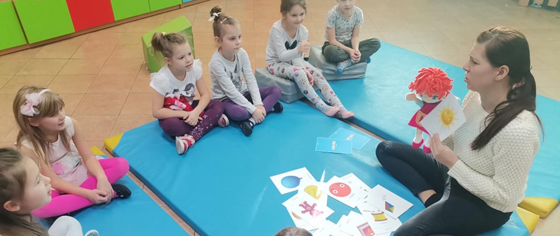 Dzieci siedzą na podłodze na przeciwko kobiety trzymającej w prawym ręku lalkę