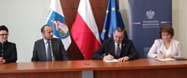 Moment podpisania umowy 21.03.2025r. od prawej Pani Skarbnik, Pan wójt w chwili podpisywania umowy w tle flagi oraz baner Wojewody Małopolskiego. 