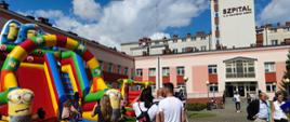 Festyn Profilaktyczno Zdrowotny w ogrodach przasnyskiego szpitala. Zdjęcie przedstawia uczestników Festynu zgromadzonych wokół atrakcji dla dzieci: dmuchanej zjeżdżalni oraz dmuchanego placu zabaw.