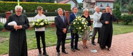 Zdjęcie grupowe zrobione na zewnątrz, na chodniku z kostki brukowej, przedstawiające sześcioosobową delegację stojącą w rzędzie. Po lewej stronie stoi ksiądz w czarnej sutannie, trzymający białą chusteczkę. Obok niego młody mężczyzna w białej koszulce polo trzyma duży wieniec z białych róż i zieleni. Następnie stoją trzej mężczyźni w garniturach (dwaj pierwsi w ciemnych, trzeci w jasnej koszuli i marynarce niesionej w ręku) – jeden z nich również trzyma duży biało-zielony wieniec. Obok nich stoi kobieta w czarnej bluzce i wzorzystej spódnicy. Po prawej stronie rząd zamyka drugi ksiądz w sutannie, wykonujący gest dłonią przy twarzy. W tle widać domy jednorodzinne, zadbane żywopłoty, drzewa iglaste oraz lekko zachmurzone niebo.