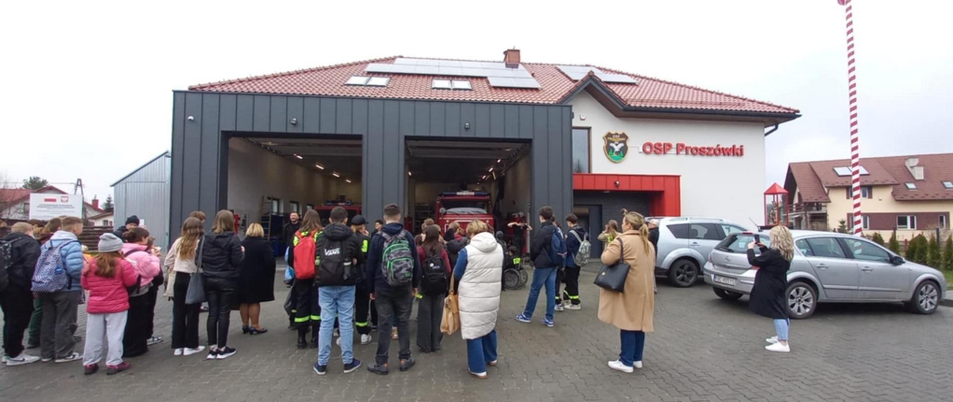 Grupa osób, widziana od tyłu, stoi przed budynkiem Ochotniczej Straży Pożarnej w Proszówkach.