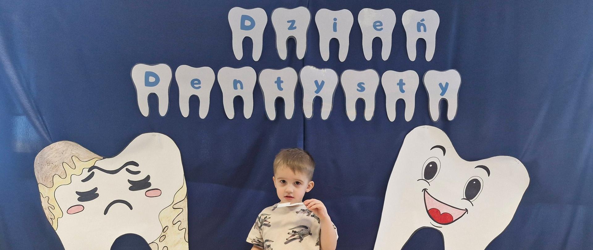 Na zdjęciu widać chłopca z grupy Delfinki, chłopczyk stoi na tle z napisem dzień dentysty oraz z rysunkiem dużego zdrowego i chorego ząbka. w rączkach trzyma filcową szczoteczkę oraz pastę. Pokazuje prawidłową technikę szczotkowania zębów