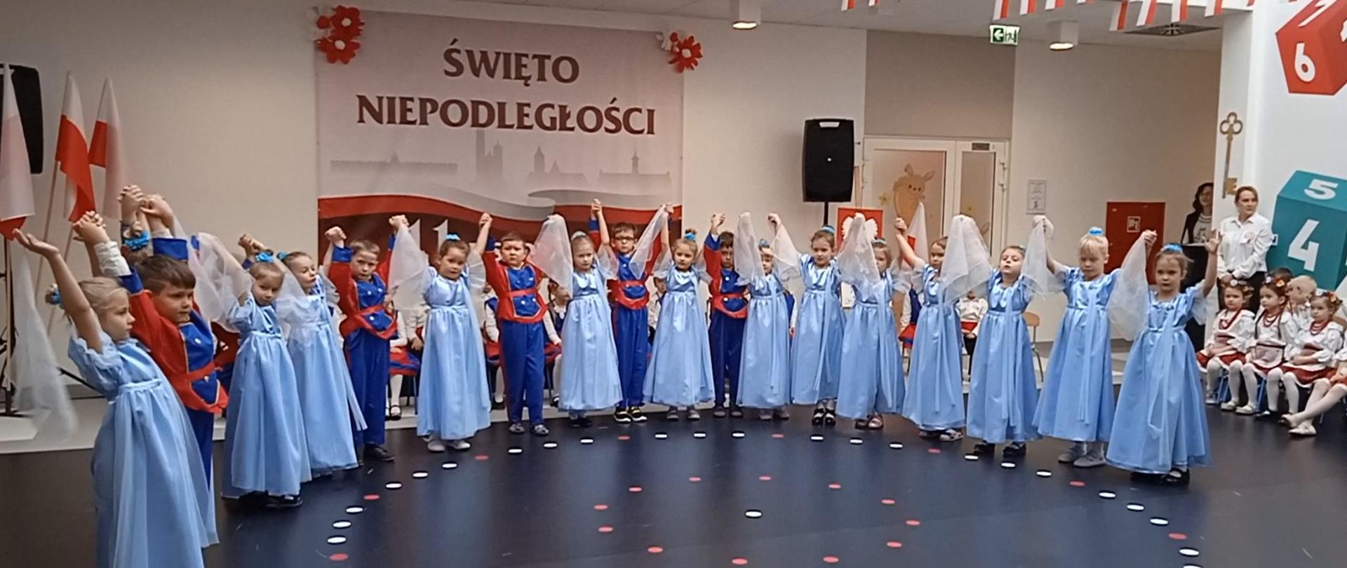 Grupa dzieci w kolorowych strojach tańczy przed banerem z napisem Święto Niepodległości
