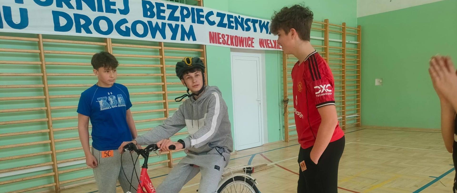 Trzech chłopców na sali gimnastycznej. Jeden z nich siedzi w kasku na rowerze a dwóch stoi obok niego.