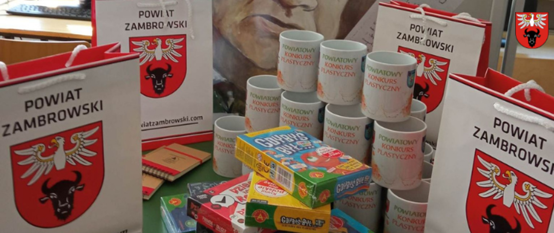na zdjęciu znajdują się nagrody ufunowane przez Starostwo Powiatowe w Zambrowie wraz z gadżetami Powiatu Zambrowskiego (widoczne są 4 torby z logo powiatu, dwa notesy z logotypami powiatu, białe kubki ustawione z napisem "POWIATOWY KONKURS PLASTYCZNY")