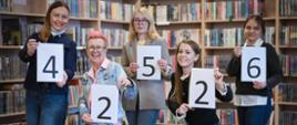 Pięć kobiet stojących w bibliotece na tle regałów z książkami trzyma kartki z cyframi 4, 2, 5, 2 i 6, układającymi się w liczbę. Uśmiechają się i pozują do zdjęcia.