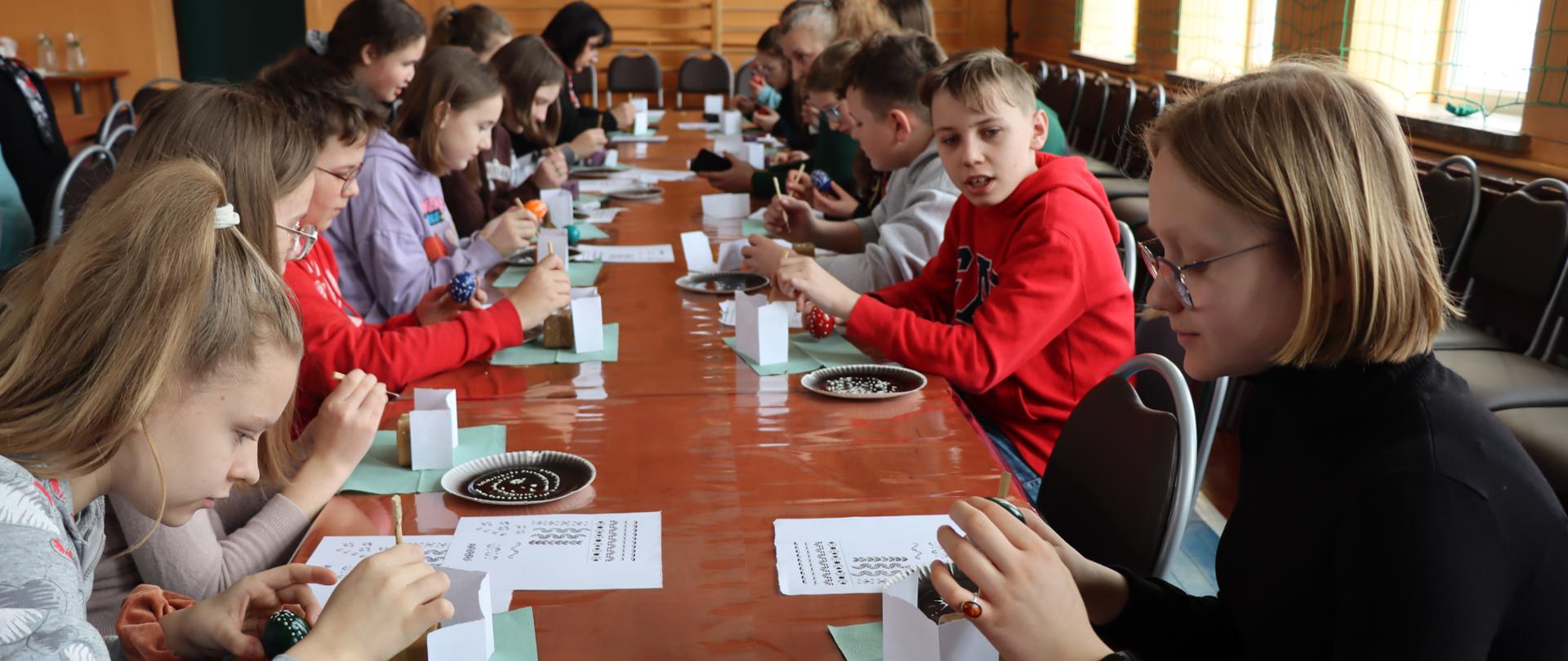 Uczniowie klasy VII Zespołu Szkół w Repkach siedzą przy stolikach, dekorując metodą butikową pisanki wielkanocne. Przed każdym uczniem stoi kominek z wgłębieniem środku,w którym znajduje się wosk podgrzewany paląca się świeczką. Uczniowie za pomocą specjalnego lejka nakładają roztopiony wosk na jajko tworząc różne wzory.