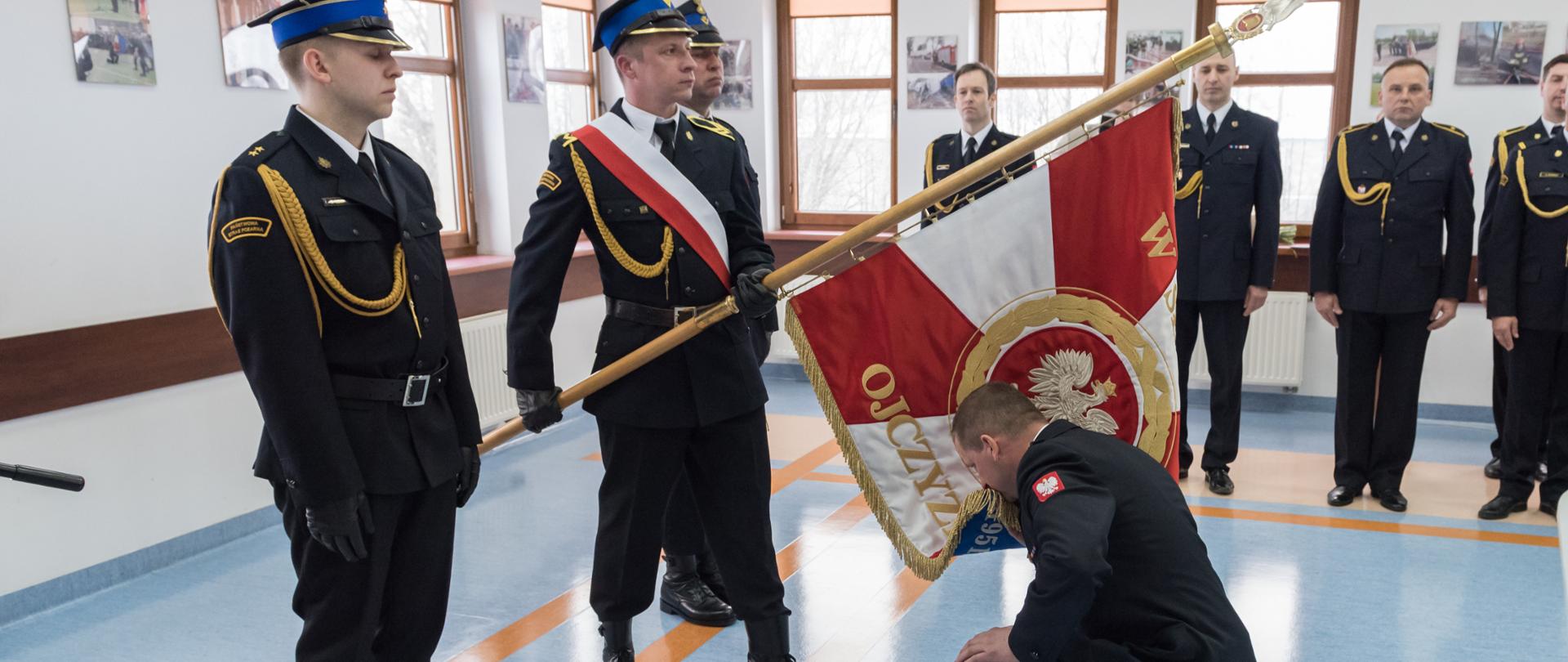 Komendant Paweł Dmowski klęczy przed pocztem sztandarowym złożonym z trzech strażaków i składa pocałunek na sztandarze Komendy Powiatowej Państwowej Straży Pożarnej w Sokołowie Podlaskim.