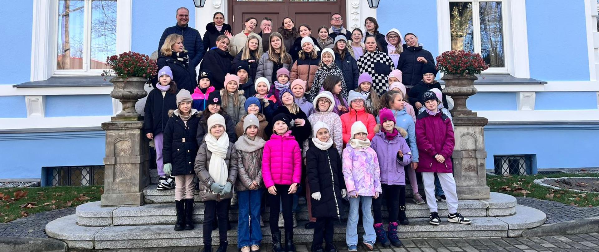 Grupa dzieci z opiekunami pozuje do zdjęcia na schodach niebieskiego budynku