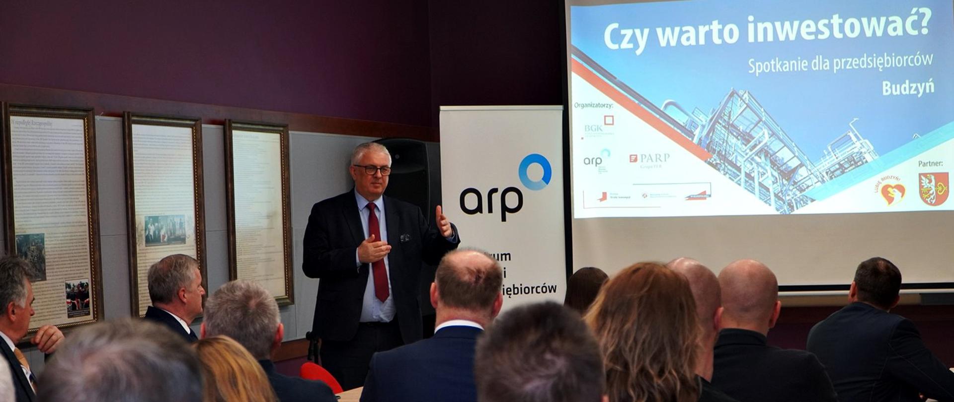 Na pierwszym palnie grupa osób siedząca przy stole, w tle przemawiający Poseł Piechowiak, na ścianie wyświetlana informacja o spotkaniu