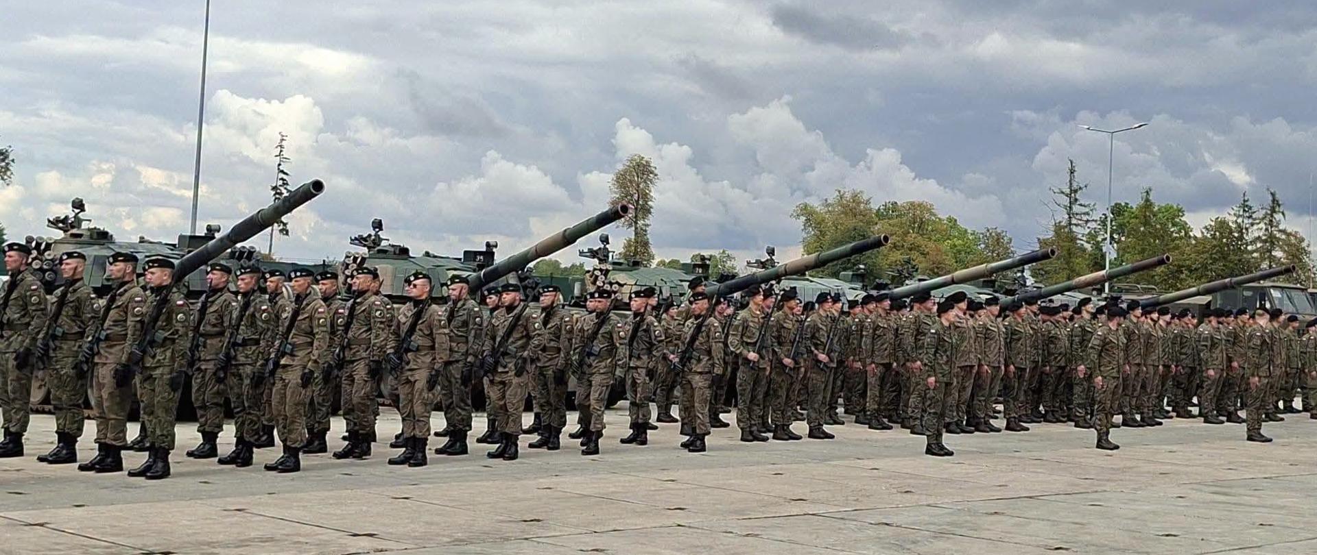 Formacja żołnierzy w mundurach wojskowych ustawionych w szeregu, z czołgami w tle pod pochmurnym niebem.
