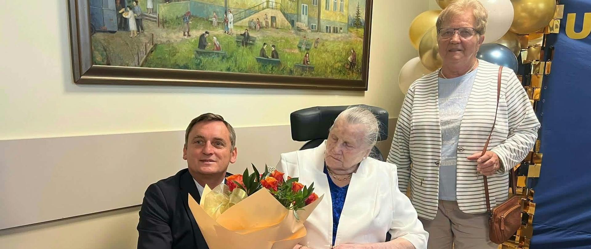 Mężczyzna w garniturze wręcza bukiet róż stuletniej jubilatce siedzącej na wózku inwalidzkim. Obok stoi uśmiechnięta kobieta w jasnym kardiganie. W tle widać dekorację z balonów oraz obraz na ścianie.