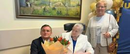Mężczyzna w garniturze wręcza bukiet róż stuletniej jubilatce siedzącej na wózku inwalidzkim. Obok stoi uśmiechnięta kobieta w jasnym kardiganie. W tle widać dekorację z balonów oraz obraz na ścianie.