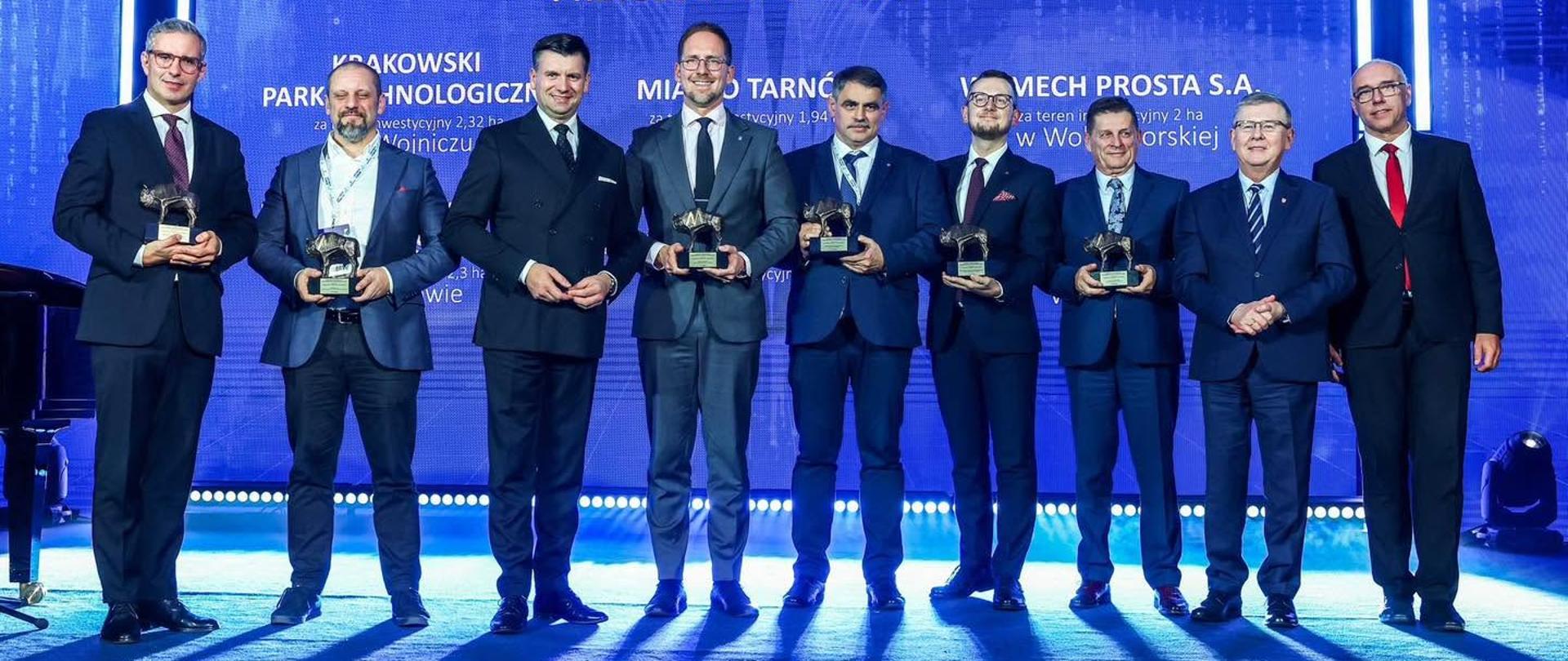 Zdjęcie grupowe wykonane podczas uroczystej gali wręczenia Nagród Gospodarczych Województwa Małopolskiego 2026, odbywającej się 27 października w Krynicy-Zdroju w ramach Krynica Forum 2026. Na scenie stoi dziewięciu mężczyzn w eleganckich garniturach, w tym przedstawiciele władz województwa oraz laureaci nagród. Część uczestników trzyma w dłoniach pamiątkowe statuetki. Wśród nagrodzonych znajduje się Burmistrz Miasta Kalwarii Zebrzydowskiej Tadeusz Stela, reprezentujący gminę wyróżnioną za teren inwestycyjny przy ul. Jagiellońskiej. W tle widoczny jest duży, niebieski ekran z napisem „Nagroda Gospodarcza Województwa Małopolskiego 2026” oraz nazwą kategorii „Małopolska Inwestycja Przyszłości – Laureaci”.