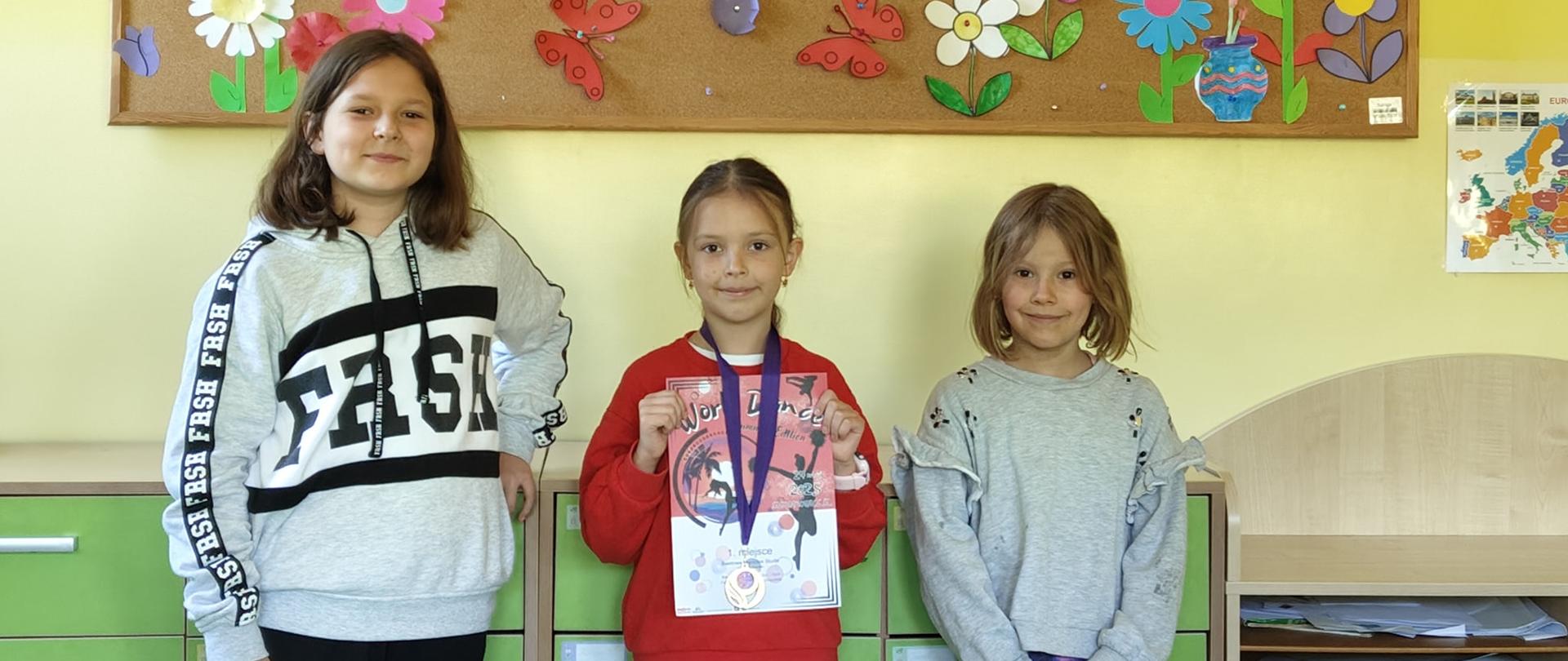 Trzy dziewczynki realizujące się jako tancerki stoją w klasie pod gazetką przedstawiającą oznaki wiosny, dziewczynka w środku trzyma dyplom za zajęcie 1. miejsca, na szyi zawieszony ma medal