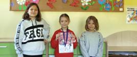 Trzy dziewczynki realizujące się jako tancerki stoją w klasie pod gazetką przedstawiającą oznaki wiosny, dziewczynka w środku trzyma dyplom za zajęcie 1. miejsca, na szyi zawieszony ma medal