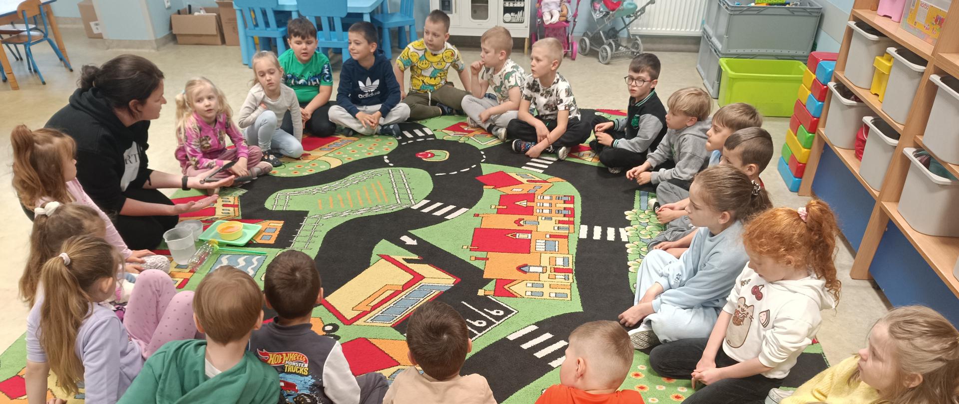 Grupa dzieci w wieku przedszkolnym siedzi na kolorowym dywanie w kole, miedzy nimi siedzi kobieta ubrana na czarno, czarne włosy. Na wprost widać niebieski mały stolik i krzesła w tym samym kolorze, obok mała kuchenka do zabawy, 2 wózki z lalkami, skrzynie z klockami. Po prawej stronie półki z pojemnikami. 