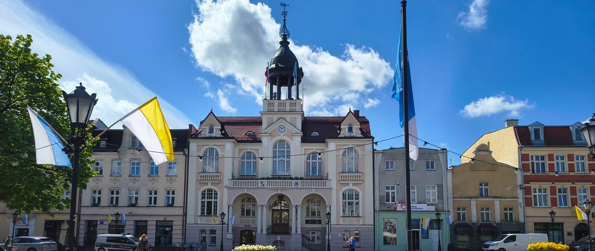 Na górze zdjęcia błękitne niebo na tle którego widoczne są białe chmury. Na środku zdjęcia biały budynek z wieżyczką na środku oraz czerwonym dachem. Z prawej strony znajduje się żółty budynek, zaś z lewej zielone drzewo. W lewym dolnym rogu latarnia, obok której widnieją dwie flagi, w kolorach biało żółtym oraz biało błękitnym. W prawym dolnym rogu kwiaty w kolorze żółtym.
