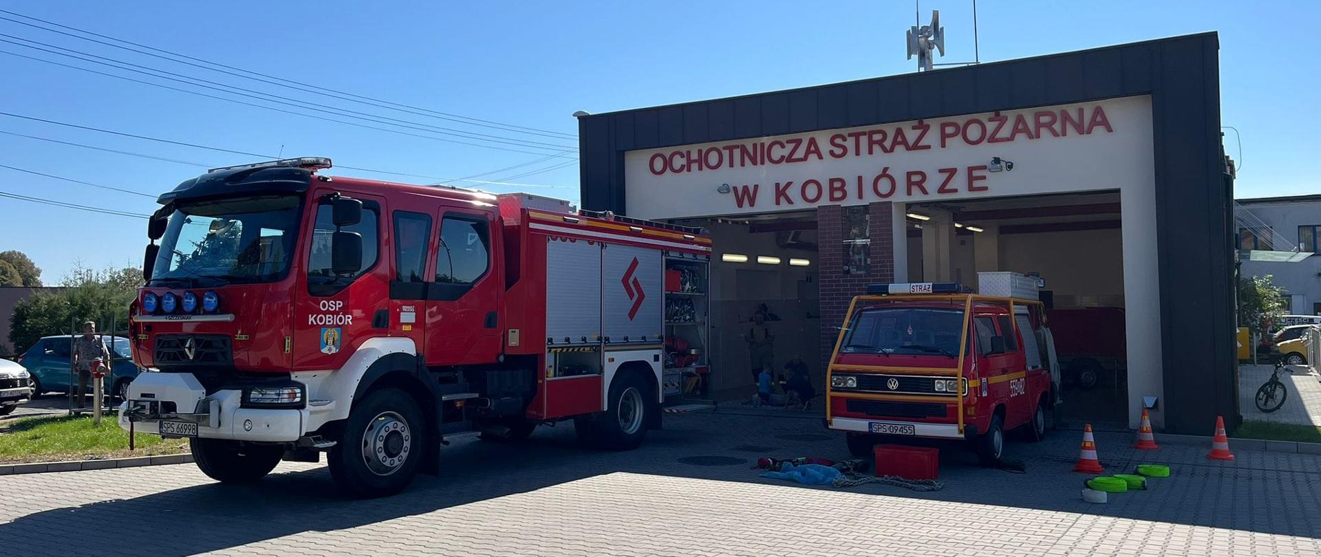 Dwa samochody strażackie stojące przed biało grafitową remizą.