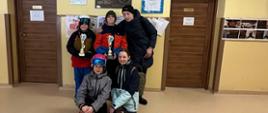 3 chłopców i dwie dziewczynki w strojach narciarskich stoją na korytarzu szkolnym pod obrazem patronki szkoły Świętej kingi, 2 chłopców trzyma w ręku złote puchary