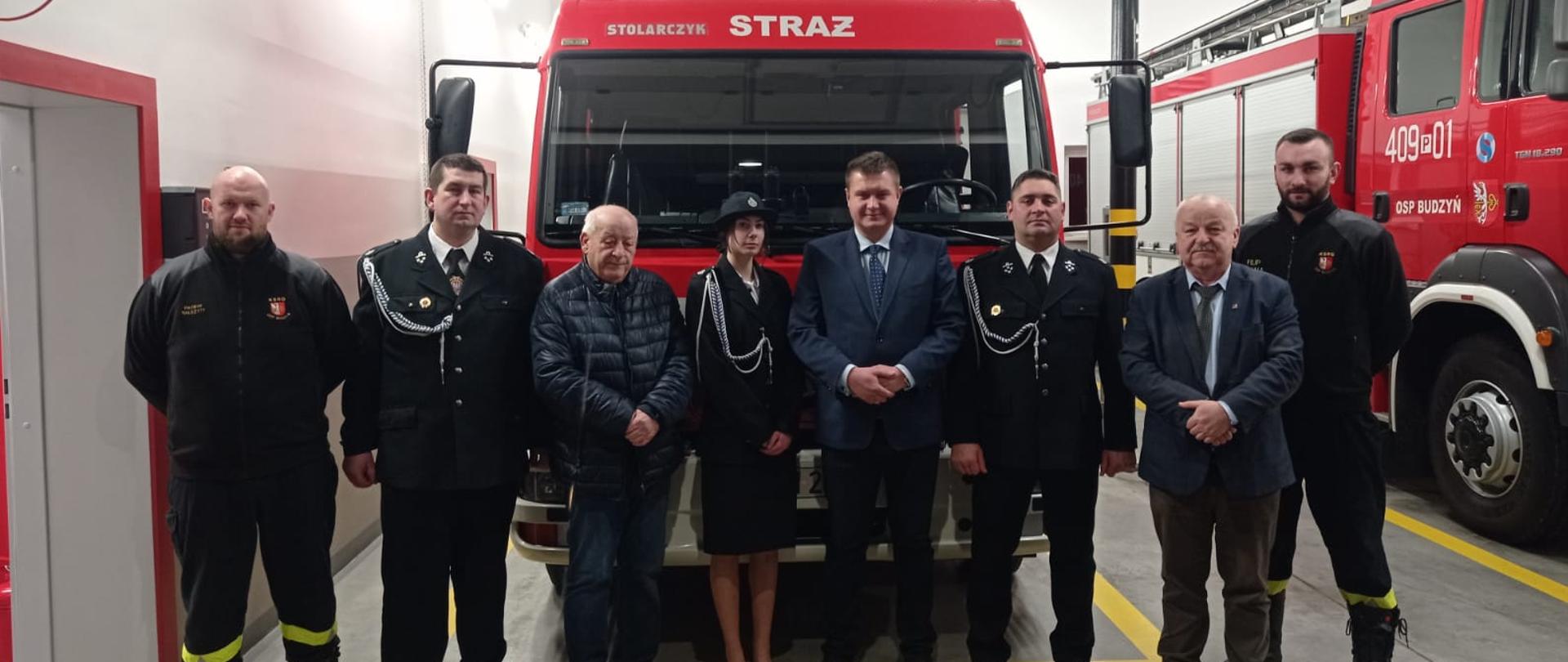 Na zdjęciu widzimy 8 osób stojących obok siebie, za nimi duży samochód ratowniczy gaśniczy.