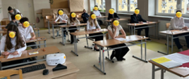 Sala lekcyjna, na niej rozmieszczone ławki szkolne przy których siedzą elegancko ubrani uczniowie. Ich twarze są zasłonięte uśmiechniętymi minkami. Z tyłu po lewej stronie widać nachylającą się nad dzieckiem panią. W tle po lewej stronie widać drzwi, a po prawej okna.