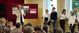 Pani dyrektor przemawia do mikrofonu, za nią stoi na drewnianych nogach gazetka z trzema plakatami przedstawiającymi chrzest Polski, obok stoi czworo uczniów występujących w akademii