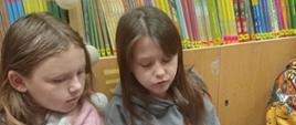 Dwie dziewczynki z klasy 3 siedzą na piankowych matach w bibliotece i czytają głośno fragmenty książki